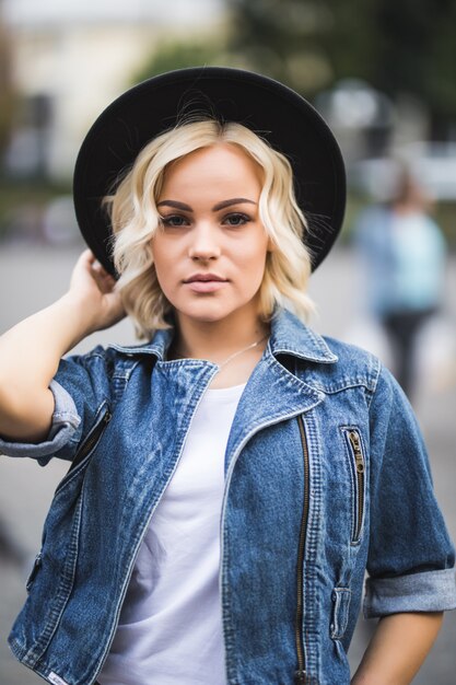 Portrait of serious blonde girl in city centre dressed up in jeans suite and white t-shirt and black hat