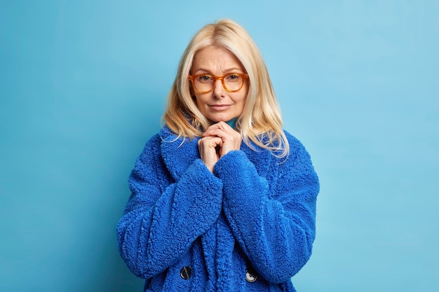 Portrait of serious blonde woman keeps hands pressed together near chin wears optical glasses and fur coat has mysterious look.