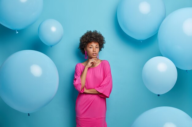 真面目な自信のある女性の肖像画は、あごを持って直接見て、バラ色のドレスを着て、青い壁で隔離されたパーティーにいる風船に対してポーズをとります。エレガントな服を着た魅力的な女性モデル