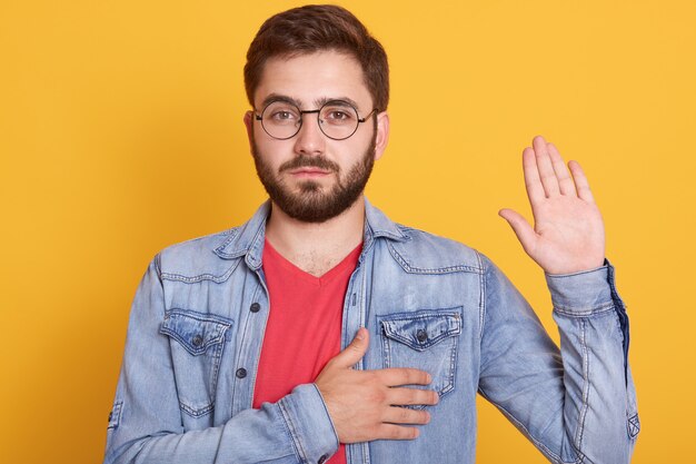 Portrait of serious confident magnetic young man looking directly  raising one hand, putting one hand to heart, making oath