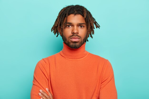 Portrait of serious confident man has healthy dark skin, dreads and beard, looks straightly at camera, has pensive expression