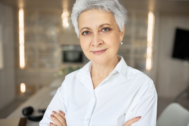 Portrait of serious confident middle aged woman with gray short hair, green eyes, wrinkles and charming smile posing indoors with arms folded