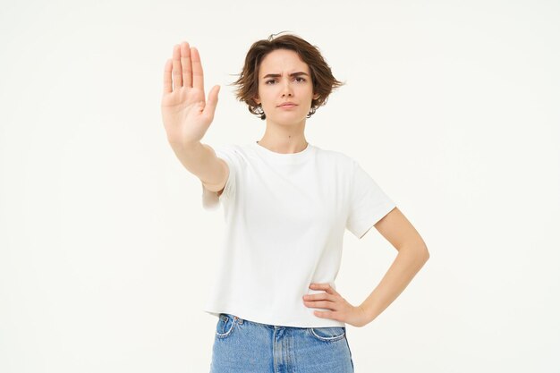 Portrait of serious confident young woman extends one hand shows stop disapproval gesture looking