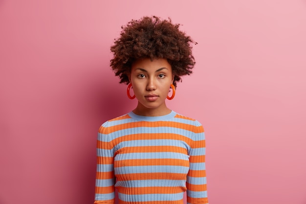 Foto gratuita ritratto di bella giovane donna dai capelli riccia seria con espressione del viso calmo, ha un aspetto fiducioso, vestito con un maglione a righe a maniche lunghe, isolato sul muro rosa. nessuna emozione