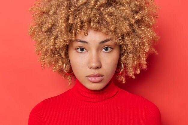 Portrait of serious curly haired woman looks attentively at camera has calm expression healthy skin natural beauty wears casual turtleneck isolated over vivid red background listens interlocutor