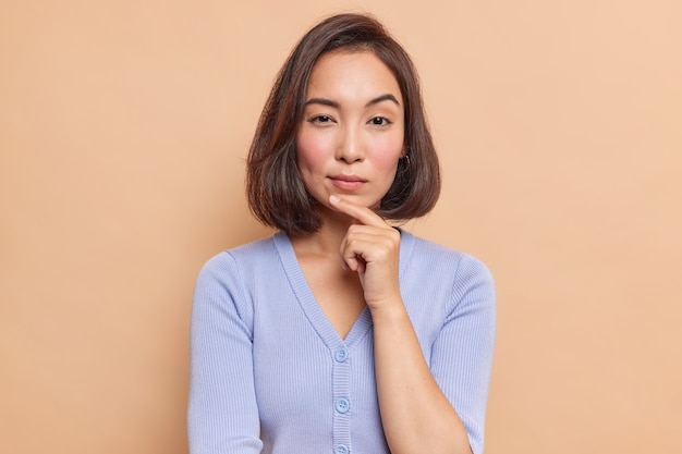 Портрет серьезной темноволосой азиатской женщины, держащей палец на подбородке, загадочно смотрит спереди и рассматривает что-то в синем джемпере, изолированное над коричневой стеной