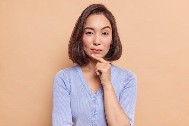 Portrait of serious dark haired Asian woman keeps finger on chin looks mysteriously at front considers something dressed in blue jumper isolated over brown wall