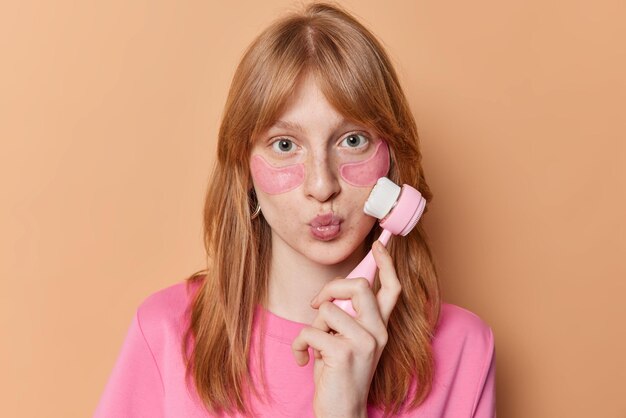 Portrait of serious European girl with red hair has freckled skin uses facial brushes applies moisturising collagen patches undergoes beauty procedures isolated over brown background Skin care