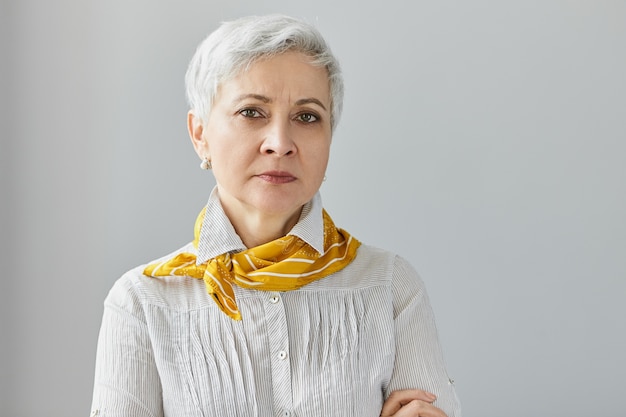 Free photo portrait of serious grandmother with wrinkles and short gray hair posing in closed posture with arms crossed, having strict look, frowning, displeased with misbehavior of her grandchildren