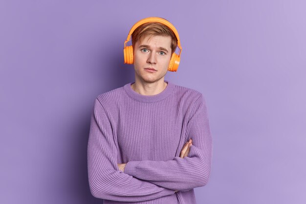 Portrait of serious handsome teenager boy stands with crossed arms and looks at camera wears headphones and jumper poses 