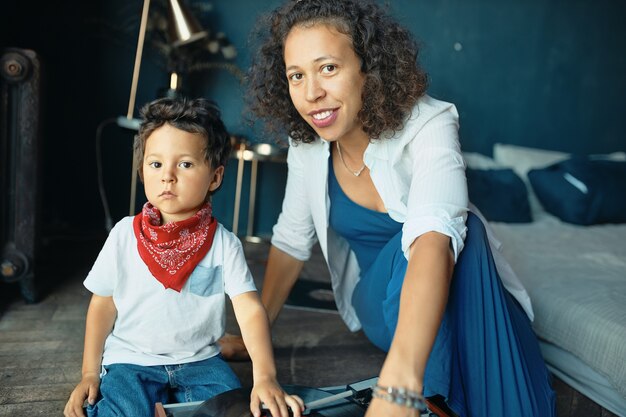 Portrait of serious little boy with chubby cheeks and red kerchief around his neck sitting on floor with mom