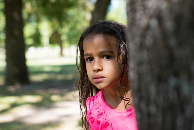 Portrait of serious little girl in park. Dark-haired girl peeking out from tree, looking at camera. Family, love, childhood concept