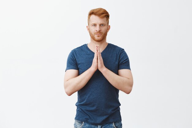 Portrait of serious-looking handsome adult redhead with muscles, holding hands in pray, making namaste gesture or bowing in asian style, staring strict over grey wall