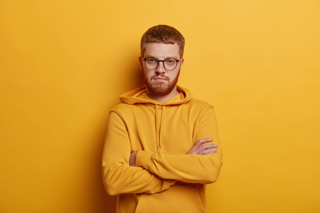 Portrait of serious looking man stands in confident gesture, keeps arms folded, has optical glasses and ginger beard, wears hoodie, poses against yellow wall. has assertive expression.