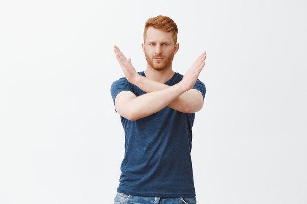 Portrait of serious-looking mature attractive redhead male with bristle in blue t-shirt, making cross with hands near chest, showing stop, enough or refusal gesture