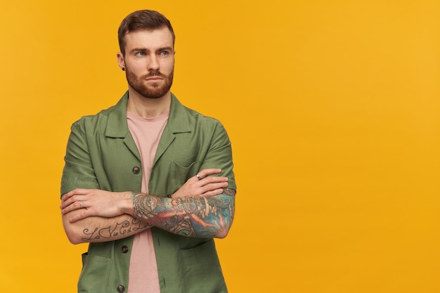 Portrait of serious male with brunette hair and beard. wearing green short sleeves jacket. has tattoo. holds arms crossed. watching to the right at copy space, isolated over yellow wall