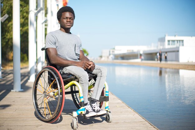 Портрет серьезного мужчины в инвалидной коляске. Афроамериканец в повседневной одежде на набережной, щурясь. Голубое небо и флаги на заднем плане. Портрет, красота, концепция счастья