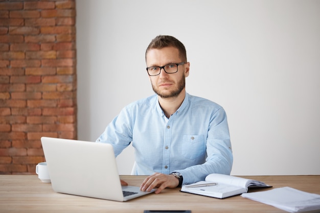 Портрет серьезного зрелого небритого бизнесмена в очках и синей рубашке, сидящей за столом, работающем над портативным компьютером, записывая задачи в тетради с расслабленным выражением.