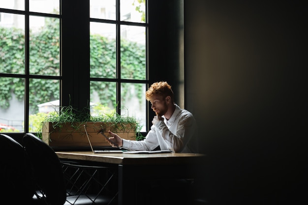 Portrait of serious redhead bearded business man holding smartphone, sitting at workplace