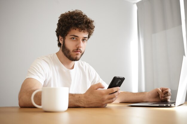 Portrait of serious stylish young man with stubble holding mobile phone dialing his friend while surfing internet on generic laptop, having hot drink at wooden table indoors,