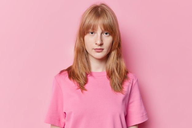 Free photo portrait of serious teenage girl with ginger hair freckles on face looks directly at camera wears casual t shirt isolated on pink background pretty redhead schoolgirl has natural beauty poses indoor