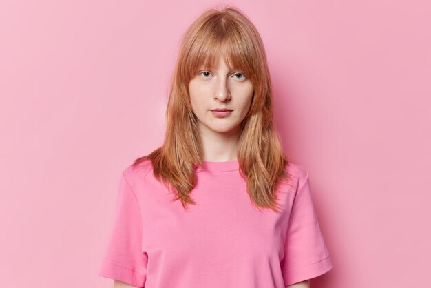 Portrait of serious teenage girl with ginger hair freckles on face looks directly at camera wears casual t shirt isolated on pink background Pretty redhead schoolgirl has natural beauty poses indoor
