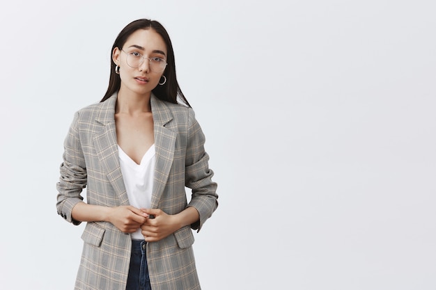 Portrait of serious unsure female entrepreneur in stylish jacket and glasses, touching fingers and gazing with doubt over grey wall