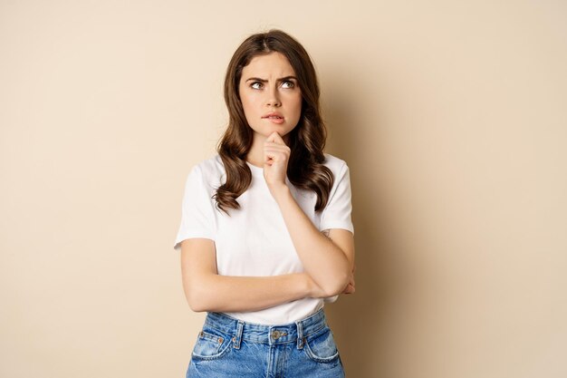 Portrait of serious woman thinking, looking up with indecisive thoughtful face, pondering, standing over beige background.