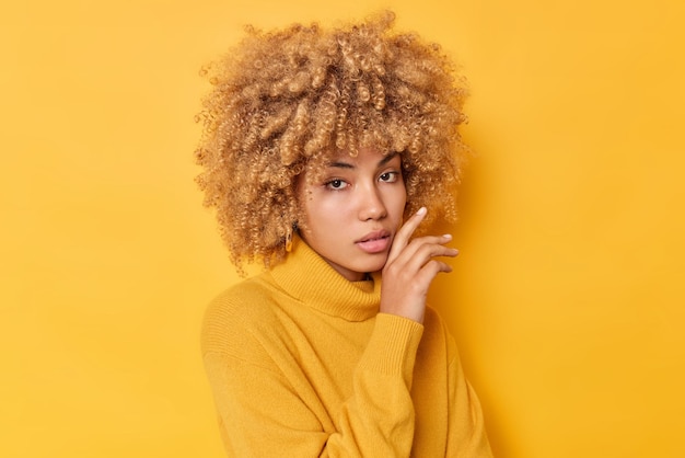 Portrait of serious woman touches gently face looks directly at camer has healthy skin curly bushy hair dressed in sweater isolated over vivid yellow background Human face expressions concept