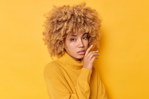 진지한 여성의 초상화가 얼굴을 부드럽게 만지며 생생한 노란색 배경 위에 격리된 스웨터를 입은 건강한 피부 곱슬곱슬한 머리카락을 가지고 있습니다. 인간의 얼굴 표현 개념