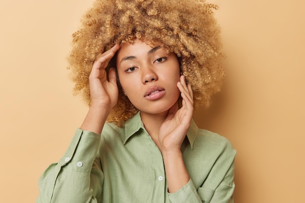 Portrait of serious woman with curly hair touches face gently has healthy clean skin looks directly at camera tenderly wears green shirt poses against brown background Natural beauty concept