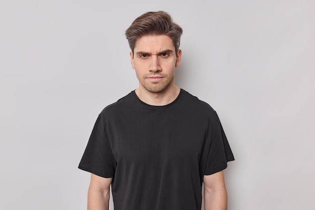 Free photo portrait of serious young man looks with strict expression dressed casually frowns face has sulking expression looks angrily at you wears casual black t shirt isolated over white background.