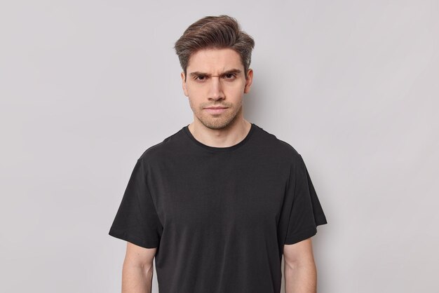 Portrait of serious young man looks with strict expression dressed casually frowns face has sulking expression looks angrily at you wears casual black t shirt isolated over white background.
