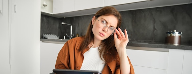 Free photo portrait of serious young woman in glasses holding tablet sitting in kitchen looking away in deep