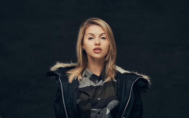 Portrait of a sexy blonde girl in a winter jacket and fleece shirt, standing in a studio. Isolated on a dark background.