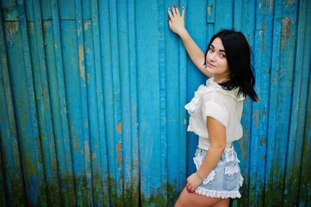 Portrait of sexy brunette girl on women's jeans shorts and white blouse against blue wooden background