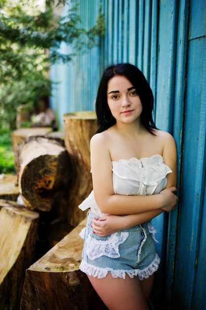 Portrait of sexy brunette girl on women's jeans shorts and white blouse against blue wooden house with stumps