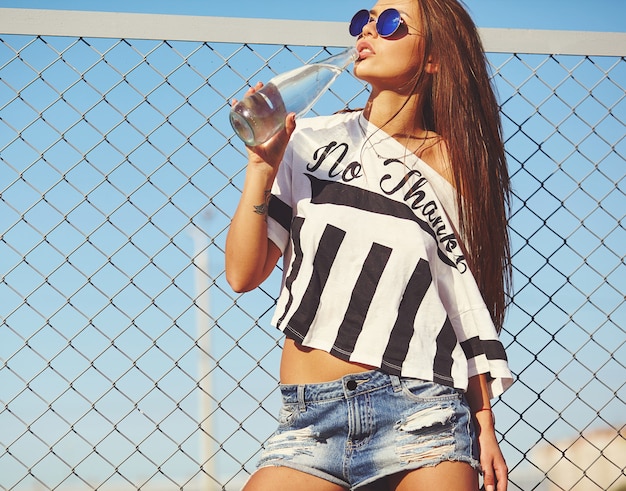 Free photo portrait of sexy glamor stylish  beautiful young woman model in bright hipster summer casual clothes posing in the street behind iron grating and blue sky. drinking water from bottle