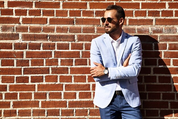 Portrait of sexy handsome fashion businessman model dressed in elegant blue suit posing near brick wall on the street background. Metrosexual