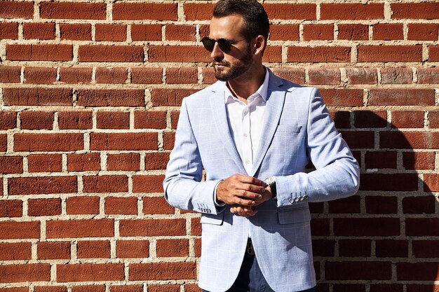 Portrait of sexy handsome fashion businessman model dressed in elegant blue suit posing near brick wall on the street background. Metrosexual