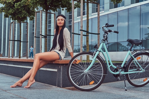 Portrait of a sexy smiling hippie female wearing blouse and shorts in a headband, sits barefoot on a bench near city bike against a skyscraper.