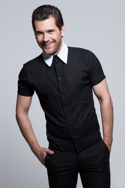 Portrait of sexy young happy man in black shirt with hands in pockets poses over gray wall.