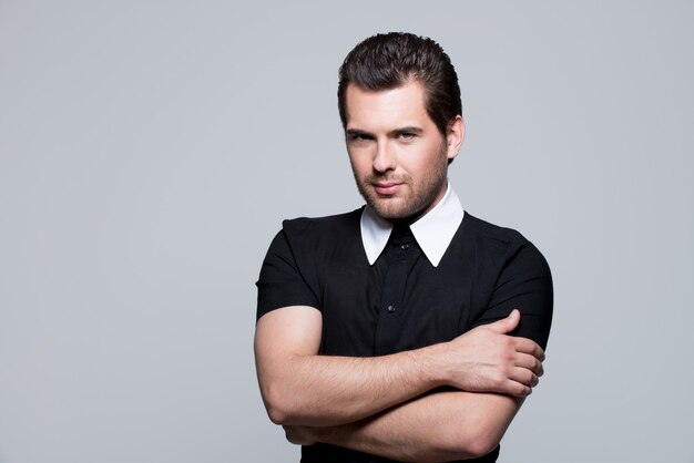 Portrait of sexy young man in black shirt with crossed arms poses over gray wall.