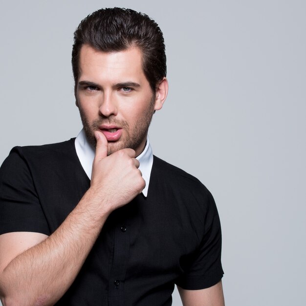 Portrait of sexy young man in black shirt with hand near face poses over gray wall.