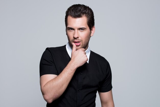 Portrait of sexy young man in black shirt with hand near face poses over gray wall.