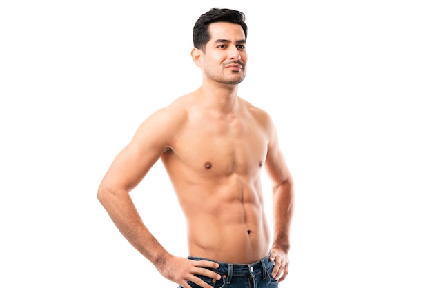Portrait of shirtless male model with muscular body standing in studio with his hands on hips