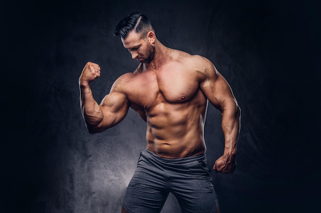 Ritratto di un maschio enorme alto senza maglietta con un corpo muscoloso con un taglio di capelli alla moda e la barba, in pantaloncini sportivi, in posa in uno studio. isolato su uno sfondo scuro.