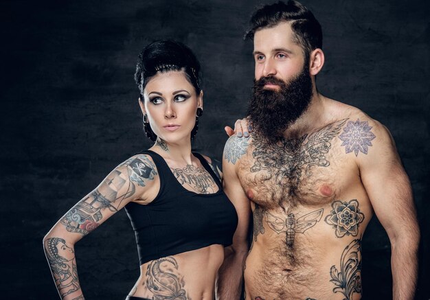 Portrait of shirtless, tattooed bearded hipster male and brunette female with tattoo ink on her torso over dark grey background.