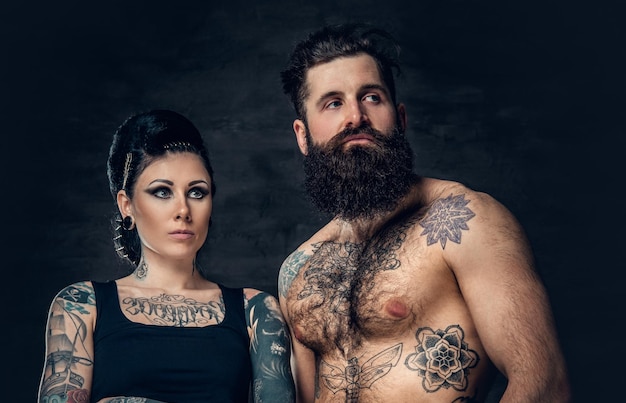 Portrait of shirtless, tattooed bearded hipster male and brunette female with tattoo ink on her torso over dark grey background.