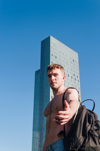 Portrait of a shirtless young man carrying backpack on shoulder giving his hand in front of camera